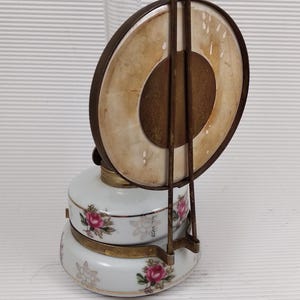 May include: Antique oil lamp with a white porcelain base decorated with pink roses and green leaves. The lamp has a clear glass chimney and a metal handle. The lamp's circular shade is a light tan color. The date 2025/4/27 00:40 is printed on the bottom.