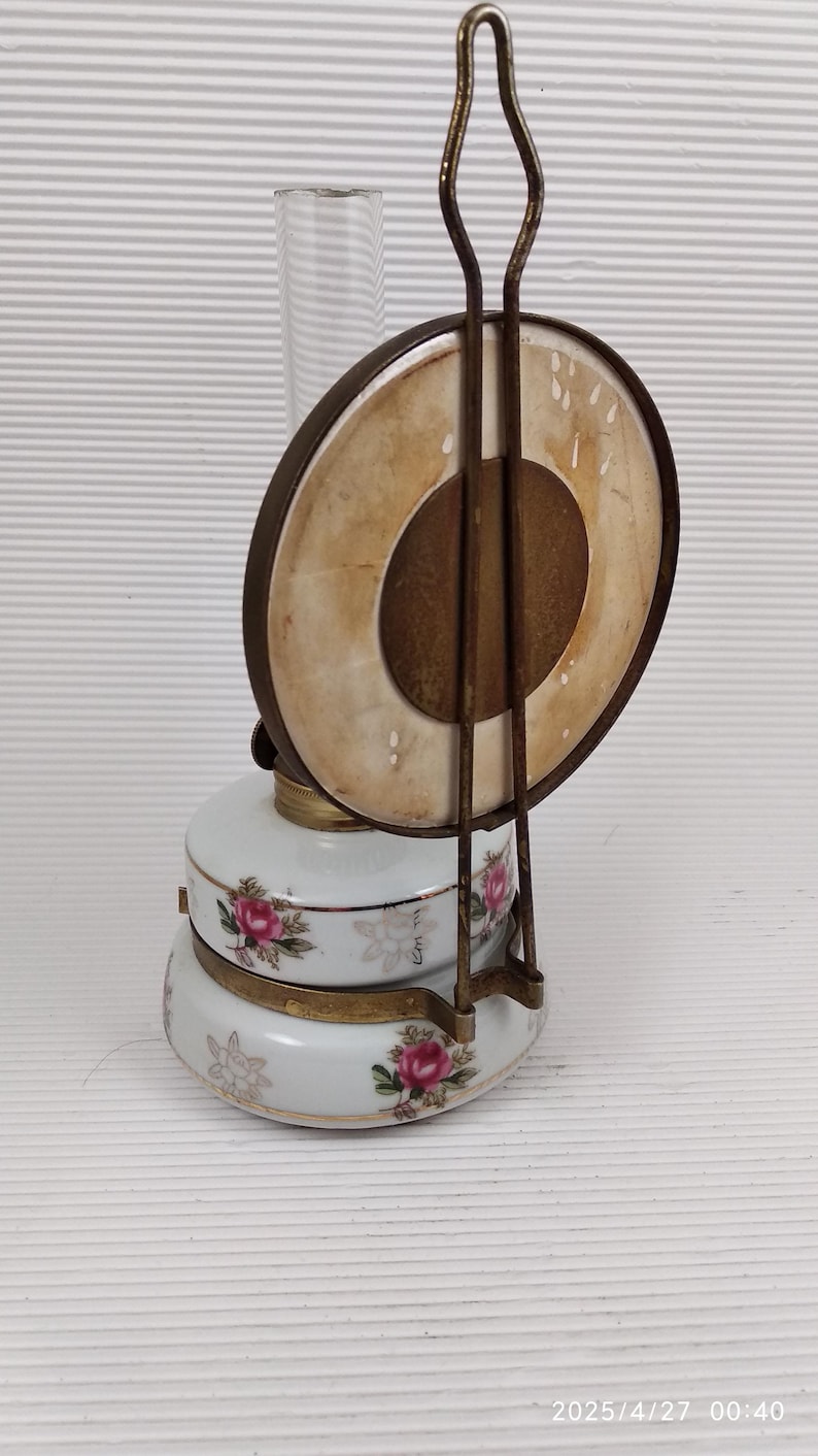 May include: Antique oil lamp with a white porcelain base decorated with pink roses and green leaves. The lamp has a clear glass chimney and a metal handle. The lamp's circular shade is a light tan color. The date 2025/4/27 00:40 is printed on the bottom.