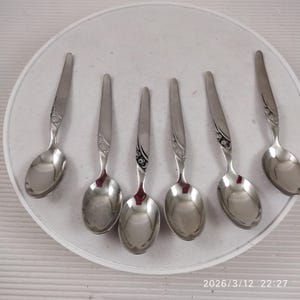Könnte beinhalten: Sechs Edelstahl-Löffel auf einem weißen Teller. Die Löffel haben ein dekoratives Blumendesign am Griff und eine abgerundete Schüssel. Die Löffel sind reflektierend und scheinen neu zu sein.