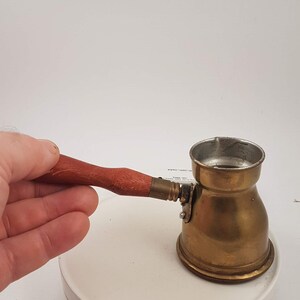 May include: A small brass coffee pot with a wooden handle. The pot has a round body and a spout on the side. The handle is attached to the pot with a metal hinge.