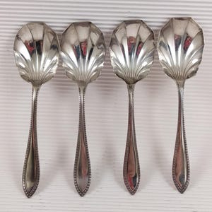 Könnte beinhalten: Vier silberne Servierlöffel mit muschelförmigen Schalen und langen, detaillierten Griffen. Die Löffel sind in einer Reihe vor einem weißen, strukturierten Hintergrund angeordnet. Die Löffel reflektieren das Licht und unterstreichen ihr aufwendiges Design und ihre polierte Oberfläche.
