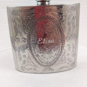 Op de afbeelding: Een zilverkleurige flacon met een gegraveerd bloemmotief en de naam "Elisa" in een ovale lijst. De flacon heeft een schroefdop en een rechthoekige vorm met afgeronde randen. De flacon is waarschijnlijk van metaal.