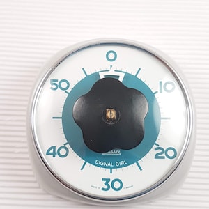 May include: A white and blue kitchen timer with a black knob. The timer is round and has a silver rim. The face of the timer has numbers from 0 to 60, and the words "Signal Girl" and "Made in Germany".