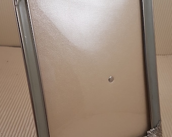 May include: A silver picture frame with a light gray mat and ornate corners. The frame is standing on a textured surface.