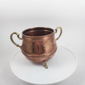 Könnte beinhalten: Eine kleine, kupferfarbene Zierschale mit zwei Messinggriffen und drei Messingfüßen. Die Schale hat einen abgerundeten Körper mit geätzten Details und einen polierten Innenraum. Die Schale steht auf einer weißen Oberfläche.