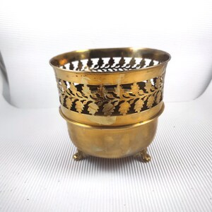 Op de afbeelding: Een vintage messing plantenbak met een decoratief opengewerkt ontwerp met bladeren en bloemen. De plantenbak heeft een afgeronde basis die wordt ondersteund door drie kleine voetjes. Het messing heeft een warme, verouderde patina.