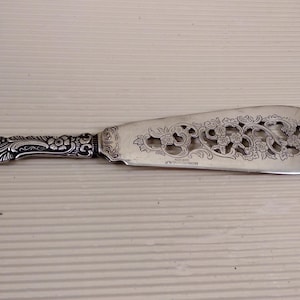 Puede incluir: Un cuchillo de mantequilla de plata con un diseño floral ornamentado en el mango y la hoja. La hoja está perforada con un patrón decorativo.