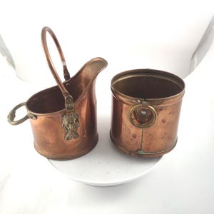 Puede incluir: Dos recipientes antiguos de cobre. Uno es una jarra pequeña con asa y una cabeza de león decorativa. El otro es un cubo pequeño con asa de anillo. Ambos tienen un color marrón rojizo cálido.