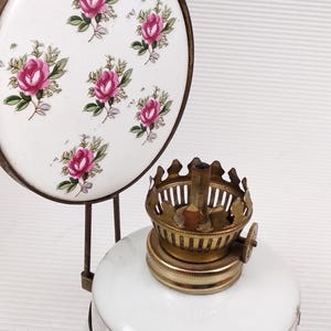 May include: Vintage oil lamp with a white ceramic base and a brass burner. The lamp features a decorative floral design with pink roses. A matching ceramic trivet with the same rose pattern is also visible.