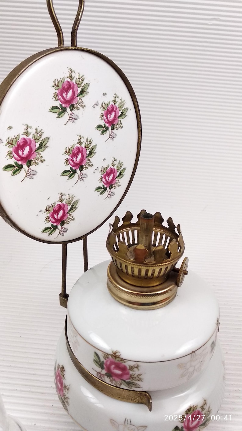 May include: Vintage oil lamp with a white ceramic base and a brass burner. The lamp features a decorative floral design with pink roses. A matching ceramic trivet with the same rose pattern is also visible.