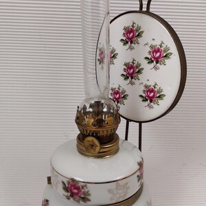 May include: A vintage white oil lamp with a clear glass chimney and brass accents. The lamp is decorated with pink rose floral patterns and has a matching decorative plate with a handle. A classic home decor item.