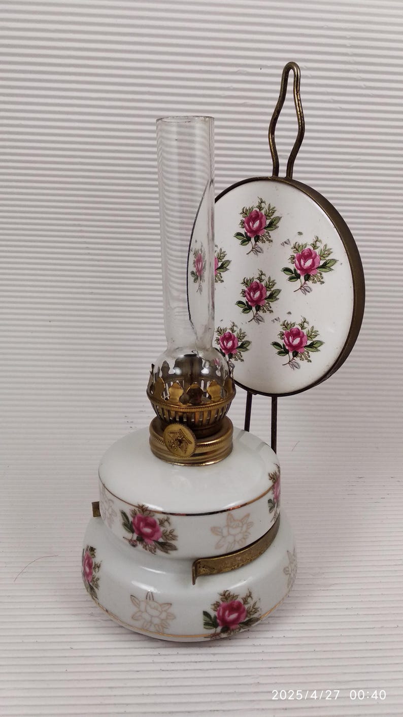 May include: A vintage white oil lamp with a clear glass chimney and brass accents. The lamp is decorated with pink rose floral patterns and has a matching decorative plate with a handle. A classic home decor item.
