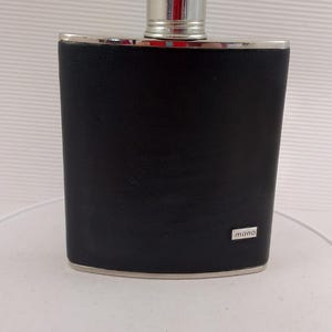 May include: A black flask with a silver cap and trim. The flask has a rectangular shape and a textured surface. The word "mano" is printed on the lower right corner. The flask is likely made of stainless steel.