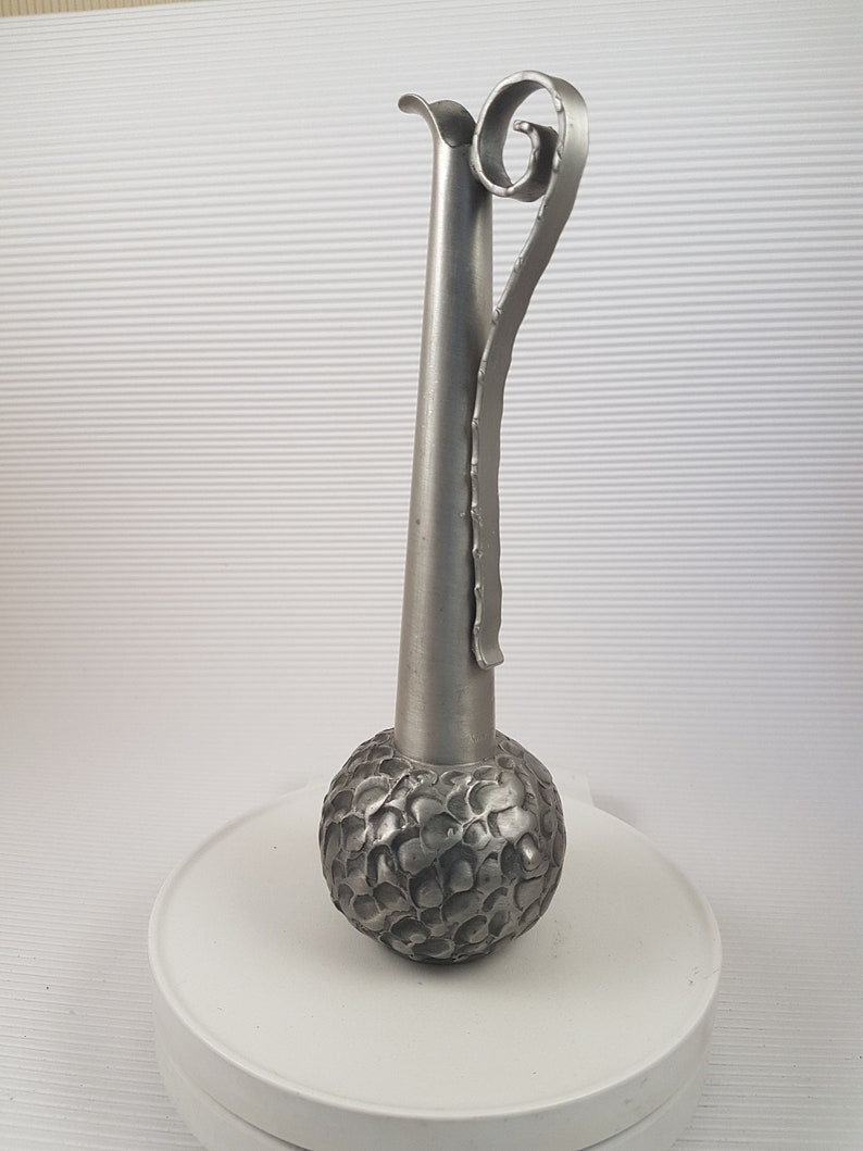 K&ouml;nnte beinhalten: Eine silberne Metallvase mit einem strukturierten, runden Boden und einem langen, schlanken Hals. Die Vase hat einen einzigartigen, gekr&auml;uselten Griff.