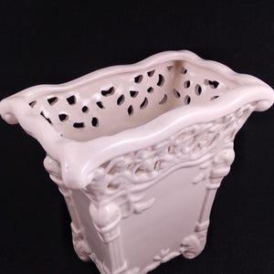 Op de afbeelding: Een rechthoekige, crèmekleurige keramische plantenbak met sierlijke details. De plantenbak heeft een geperforeerd ontwerp langs de bovenrand en decoratieve reliëfpatronen aan de zijkanten, ondersteund door kleine voetjes. De algehele stijl is klassiek en elegant.
