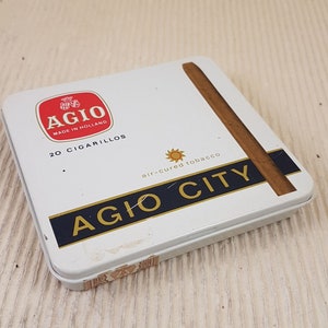 以下が含まれることがあります： 白地に赤と白のロゴ「Agio Made in Holland」が書かれた白いブリキ箱。箱には20本のシガリロが入っており、「Agio City air-cured tobacco」と書かれた黒と金の帯が付いている。