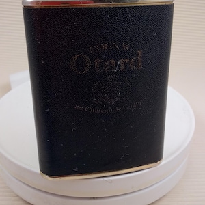 Puede incluir: Una petaca de cuero negro con ribete dorado y tapa plateada. La petaca tiene las palabras "Cognac V.S.O.P. Grand" grabadas en la parte delantera.