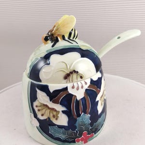 May include: A ceramic honey pot with a lid featuring a bee figurine. The pot is decorated with white flowers, green leaves, and a dark blue background. It has a small handle and a spoon-shaped extension.