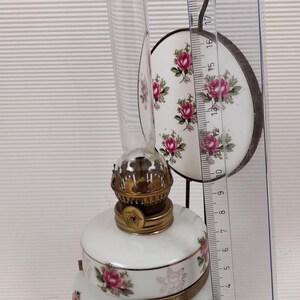May include: A white porcelain oil lamp with a clear glass chimney and a decorative floral pattern featuring pink roses. The lamp has brass accents and a round, matching shade. A ruler shows the lamp is approximately 6 inches tall.