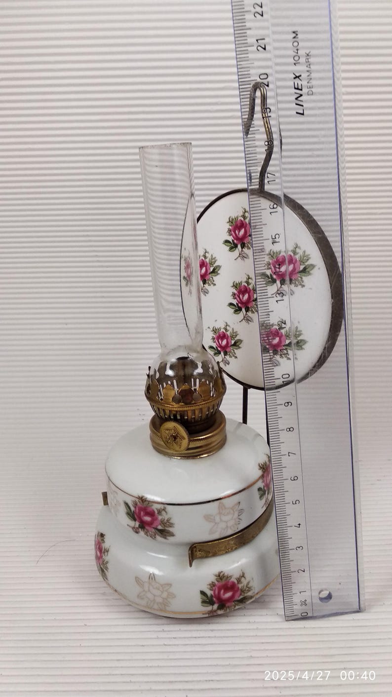 May include: A white porcelain oil lamp with a clear glass chimney and a decorative floral pattern featuring pink roses. The lamp has brass accents and a round, matching shade. A ruler shows the lamp is approximately 6 inches tall.