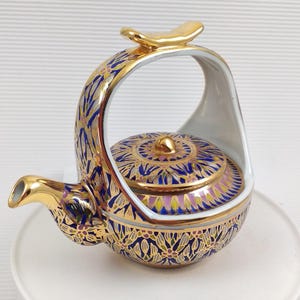 Peut inclure: Une théière décorative en céramique avec une anse et un bec dorés. La théière est ornée de motifs floraux bleus et dorés complexes. Le couvercle est également décoré d'un motif assorti. La théière repose sur une assiette blanche.