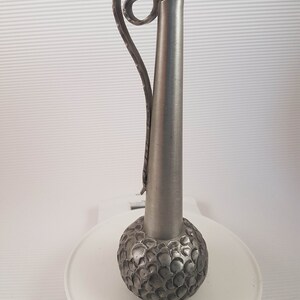 K&ouml;nnte beinhalten: Eine silberne Zinnvase mit einem strukturierten, runden Boden und einem langen, schlanken Hals. Die Vase hat einen dekorativen, gekr&auml;uselten Griff.