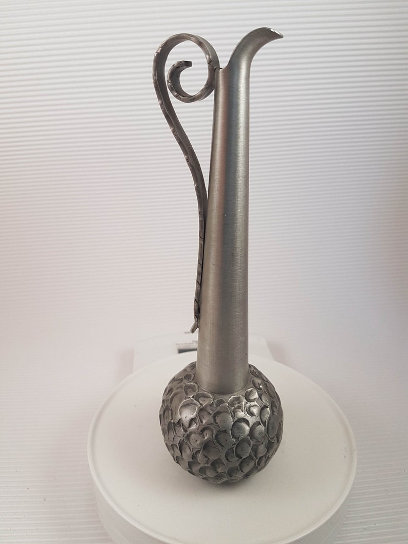 K&ouml;nnte beinhalten: Eine silberne Zinnvase mit einem strukturierten, runden Boden und einem langen, schlanken Hals. Die Vase hat einen dekorativen, gekr&auml;uselten Griff.