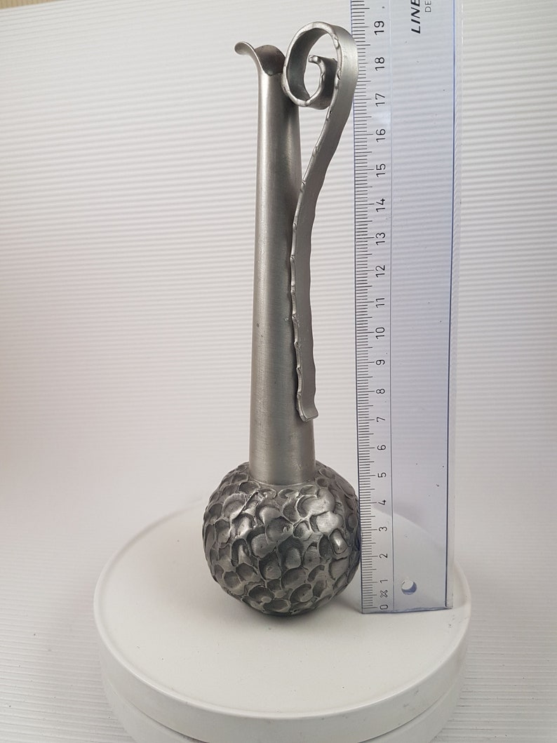 K&ouml;nnte beinhalten: Eine silberne Metallvase mit einem strukturierten, runden Boden und einem hohen, schlanken Hals. Die Vase hat einen dekorativen, gekr&auml;uselten Griff.