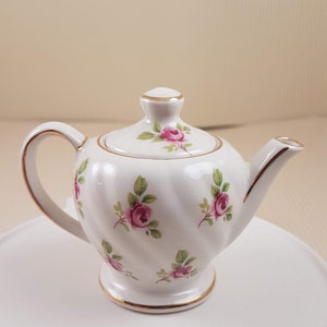 Könnte beinhalten: Eine kleine weiße Teekanne mit goldener Verzierung und einem Blumenmuster aus rosa Rosen und grünen Blättern. Die Teekanne steht auf einem weißen Sockel.