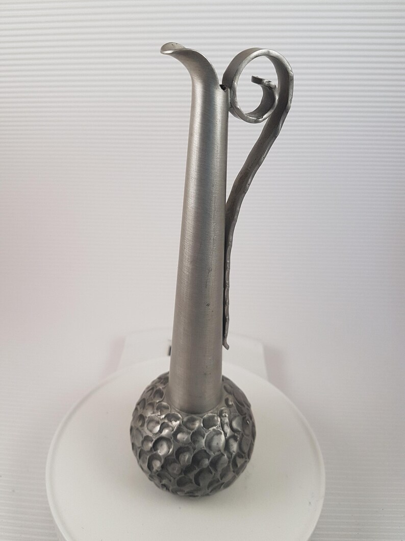 K&ouml;nnte beinhalten: Eine silberne Metallvase mit einem strukturierten, runden Boden und einem langen, schlanken Hals. Die Vase hat einen einzigartigen, gekr&auml;uselten Griff.