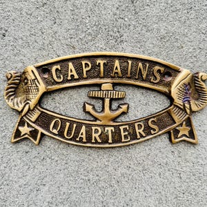 Pode incluir: Uma placa de metal de cor bronze com as palavras "CAPTAINS QUARTERS" em letras em relevo. A placa apresenta uma âncora no centro e desenhos de cabeça de elefante em cada lado. As estrelas estão em cada extremidade.