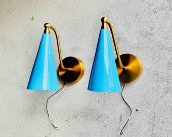 May include: A pair of wall-mounted lamps featuring bright blue conical shades and brushed gold-tone metal arms and circular wall plates. Each lamp has a curved arm connecting the shade to the base, with a thin white electrical wire extending downward.