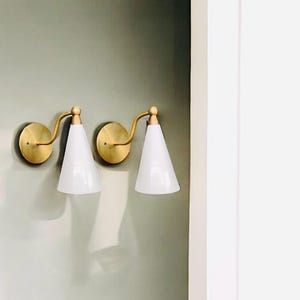 May include: Two wall sconces with brass-colored circular backplates and curved arms. Each sconce holds a white, cone-shaped shade. The sconces are mounted on a light green wall.