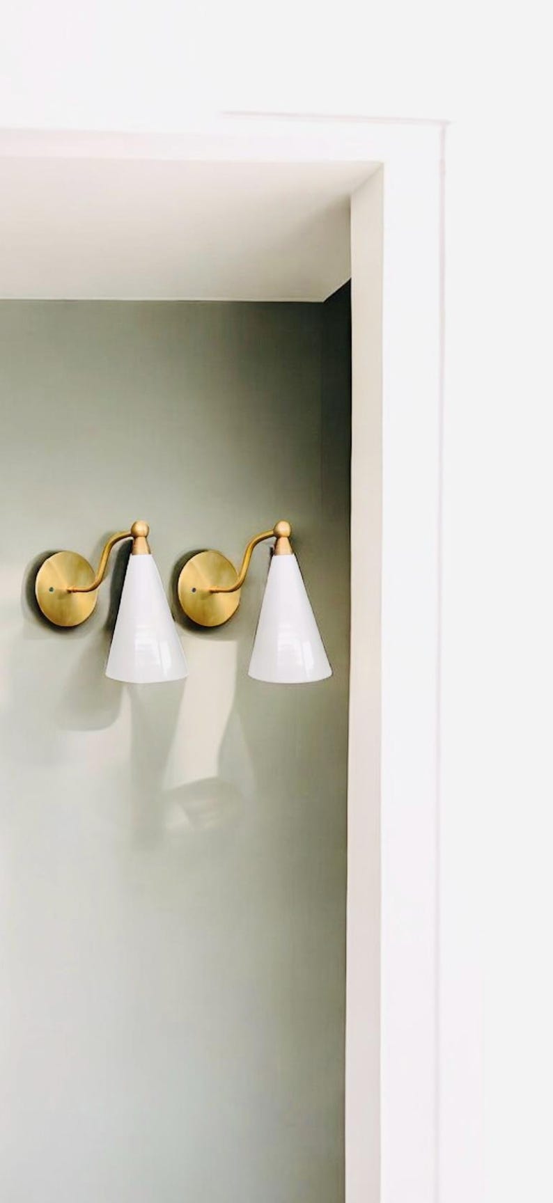 May include: Two wall sconces with brass-colored circular backplates and curved arms. Each sconce holds a white, cone-shaped shade. The sconces are mounted on a light green wall.