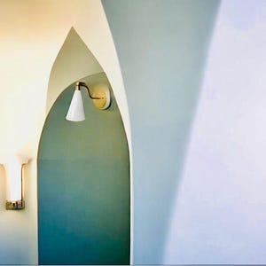 May include: A modern interior design featuring a white wall sconce with a conical shade and brass accents. The sconce is mounted on a teal wall within an arched doorway. Another wall sconce is visible on the left.