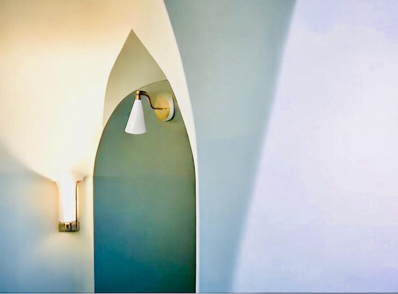 May include: A modern interior design featuring a white wall sconce with a conical shade and brass accents. The sconce is mounted on a teal wall within an arched doorway. Another wall sconce is visible on the left.