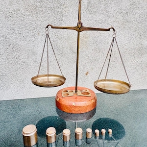 May include: A vintage brass balance scale with two circular pans hanging by chains. The scale has a wooden base and is accompanied by a set of brass weights of various sizes. The scale is likely an antique item, used for measuring.