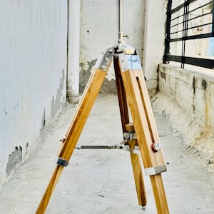 Può includere: Un treppiede in legno con dettagli in metallo e una colonna centrale regolabile in metallo. Le gambe del treppiede sono in legno chiaro e sono fissate con staffe metalliche. Il treppiede è progettato per sostenere una fotocamera o altre attrezzature.