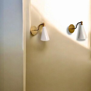 May include: Two wall-mounted sconces with a brass finish and white, cone-shaped shades. The sconces have a curved arm and a circular base. The design is minimalist and modern, suitable for interior lighting.