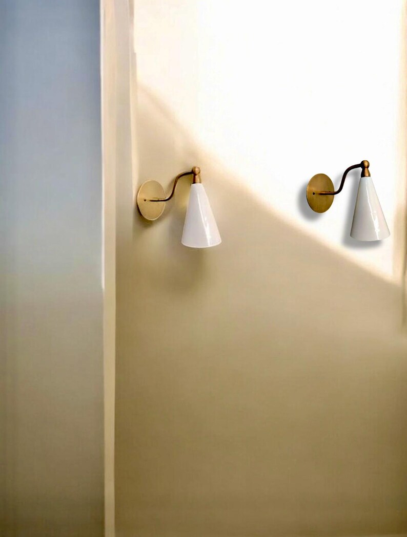 May include: Two wall-mounted sconces with a brass finish and white, cone-shaped shades. The sconces have a curved arm and a circular base. The design is minimalist and modern, suitable for interior lighting.