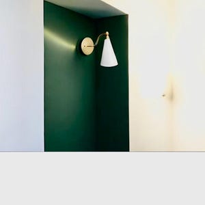 May include: A modern wall sconce with a white cone-shaped shade and a brass-colored arm and circular base. The sconce is mounted on a dark green wall, with a white wall to the side.