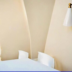 May include: A minimalist bedroom scene featuring a white bed and two white chairs against a beige wall. A brass and white sconce provides soft lighting, adding a touch of elegance to the space.