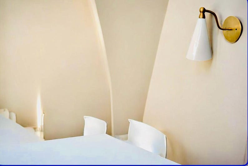 May include: A minimalist bedroom scene featuring a white bed and two white chairs against a beige wall. A brass and white sconce provides soft lighting, adding a touch of elegance to the space.
