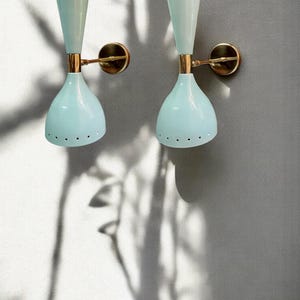 May include: Two vintage wall sconces. Each sconce has a light blue, bell-shaped shade with small holes, a gold-colored arm, and a light blue, cone-shaped top. The sconces are mounted on a gray wall.