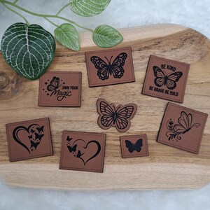 Könnte beinhalten: Eine Sammlung von beigefarbenen rechteckigen und quadratischen Aufnähern mit schwarzen, lasergravierten Schmetterlings- und Herzmotiven. Der Text umfasst "OWN YOUR MAGIC" und "BE KIND BE BRAVE BE BOLD". Die Aufnäher zeigen verschiedene Schmetterlingsmotive.