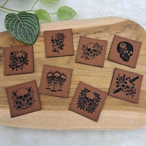 Könnte beinhalten: Acht quadratische Aufnäher aus hellbraunem Leder mit schwarzen Tintenillustrationen von Totenköpfen, Rosen und Dolchen. Die Motive zeigen unter anderem einen Totenkopf mit Rose, einen Totenkopf mit tropfender Flüssigkeit und einen Totenkopf mit Schwert.