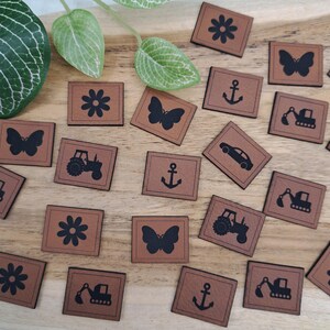 Könnte beinhalten: Eine Sammlung brauner quadratischer Kunstlederetiketten mit verschiedenen schwarzen grafischen Designs. Die Designs umfassen eine Blume, einen Schmetterling, einen Anker, ein Auto, einen Traktor und einen Bagger. Die Etiketten sind auf einer Holzoberfläche angeordnet.