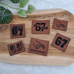 Könnte beinhalten: Eine Sammlung brauner Lederaufnäher mit schwarzem Text und Grafiken. Die Aufnäher zeigen die Zahl 67, den Satz "I WAS NOW I'M 67" sowie andere Sätze und Handgesten. Die Aufnäher sind auf einem Holztablett angeordnet.
