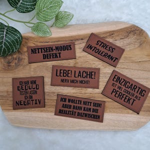 Könnte beinhalten: Braune rechteckige Aufnäher mit schwarzem Text, darunter Sätze wie "Stressintolerant" und "Einzigartig ist viel cooler als perfekt". Die Aufnäher sind auf einer Holzoberfläche angeordnet, mit grünen Blättern im Hintergrund.