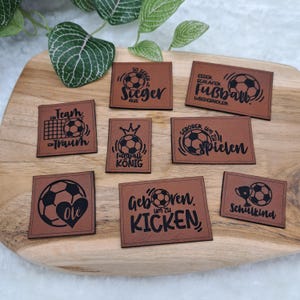 Könnte beinhalten: Eine Auswahl brauner Lederpatches mit schwarzen Fußballmotiven und Texten. Auf den Patches stehen Sätze wie "Sieger", "Fußball" und "Schulkind". Die Patches sind auf einem Holzbrett angeordnet.